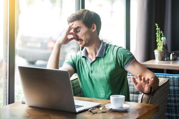 Schlechter Geruch Junger unzufriedener Geschäftsmann in grünem T-Shirt sitzt und arbeitet am Laptop und kneift sich die Nase mit negativem Emotionsgeschäft und freiberuflichem Konzept Innenaufnahme tagsüber in der Nähe des Fensters