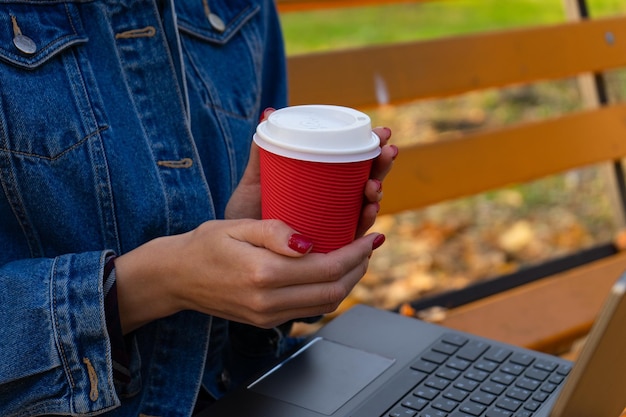 Schließen Sie bis zur Hand junge Frau, die eine Tasse Kaffee hält, während sie einen Laptop in einem Herbstpark verwendet