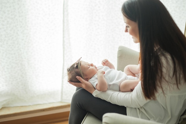 Schließen Sie das Porträt eines kleinen niedlichen dunkelhaarigen neugeborenen Mädchens in einem weißen Anzug in den Armen einer jungen Mutter Mutterschaftselternteil Gesunder Schlaf