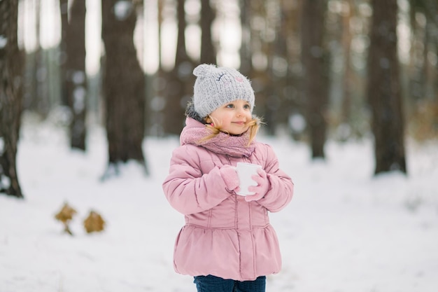 Schließen Sie das Porträt eines kleinen süßen Mädchens in rosa Kleidung mit einer Tasse heißem Tee, das draußen auf dem Hintergrund des schönen verschneiten Winterwaldes posiert und wegschaut.