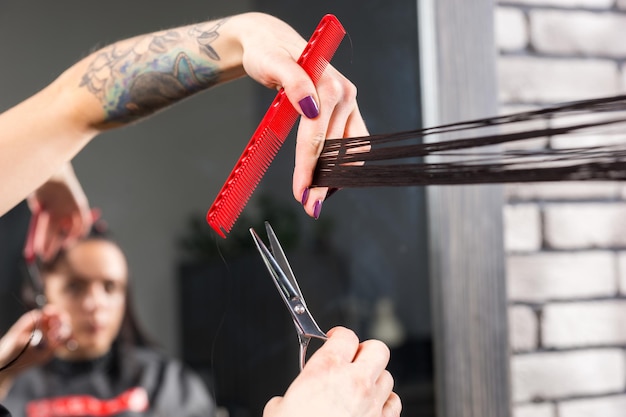 Schließen Sie die Hände eines professionellen Friseurs, der braune Haare mit einer Schere und einem Kamm einer jungen brünetten Frau schneidet und modelliert, während sie im Sessel im Schönheitssalon sitzt