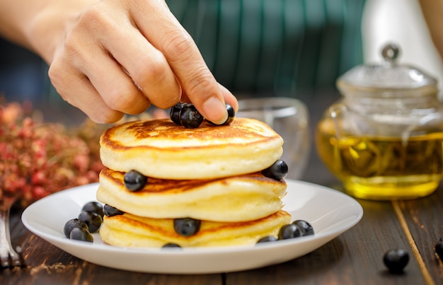 Schließen Sie die Hand der nicht erkennbaren Frau, die die Hand hält, die Pfannkuchen verziert, indem Sie Blaubeeren aufheben und den Hintergrund der Teekanne verwischen. Konzept süßes Essen.