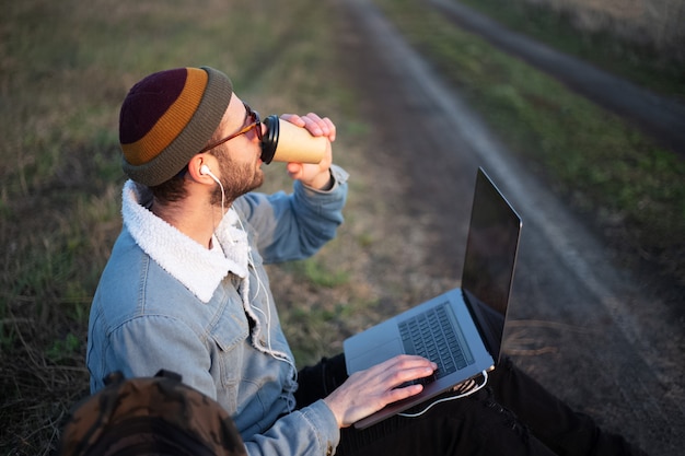 Schließen Sie herauf des jungen Mannes, der mit Laptop auf Beinen und Tasse Kaffee in der Hand sitzt