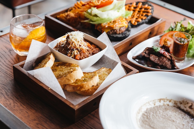 Schließen Sie herauf die Fleischsoße, die mit dem Mozzarellakäse übersteigt, gedient mit Knoblauchbrot auf Holztisch mit Holzkohlecheeseburger, gegrilltem Schweinefleisch und Cocktail.