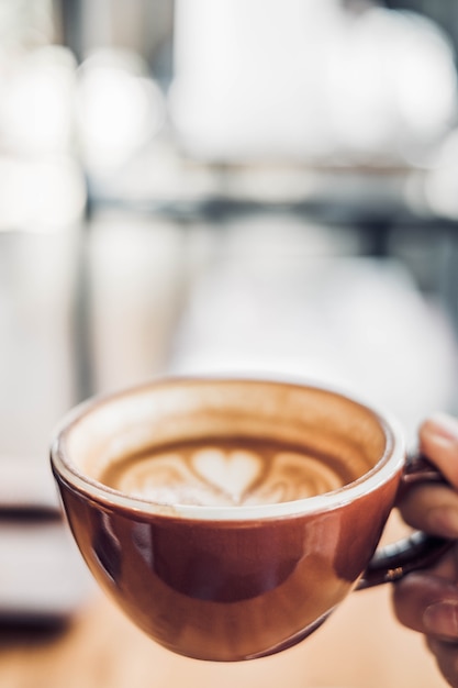 Schließen Sie herauf die Hand, die eine Schale heiße CappuccinoKaffeetasse mit Herzform Lattekunst-Unschärfecafé hält