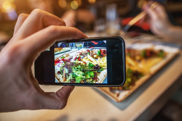 Schließen Sie herauf Fokusansicht der Hand und des Mobiles, während Sie ein Foto des Essens auf dem Teller in einem Restaurant für soziale Medien machen.