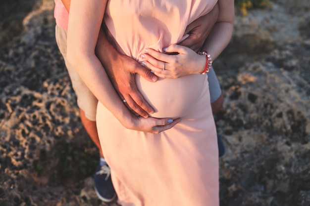 Schließen Sie herauf Foto der jungen schwangeren Frau im eleganten rosa Kleid, das ihren Bauch mit den Händen berührt