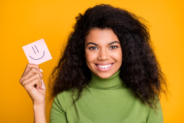 Schließen Sie herauf Foto des fröhlichen zahnigen strahlenden Mädchens, das Papier mit Emoji zeigt, das darin gemalt wird, das Emotionen lokalisierten lebendigen Farbhintergrund vergleicht