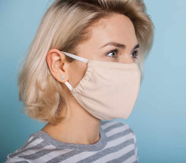 Schließen Sie herauf Foto einer reizenden blonden Dame mit medizinischer Maske auf Gesicht, das auf einer blauen Studiowand aufwirft