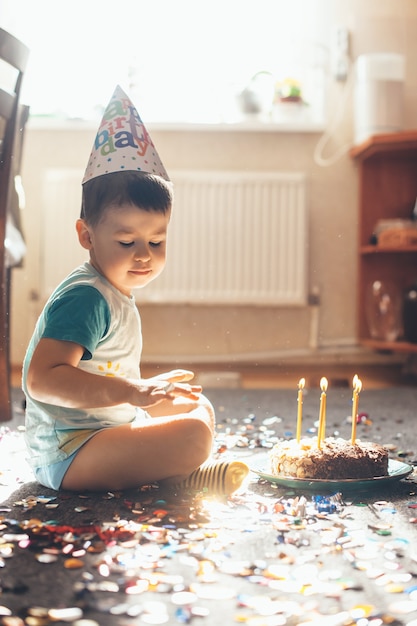 Schließen Sie herauf Foto eines kleinen kaukasischen Jungen, der seinen Geburtstag auf dem Boden feiert, der mit einem Kuchen und Konfetti aufwirft, während Sie eine Feiertagsmütze tragen