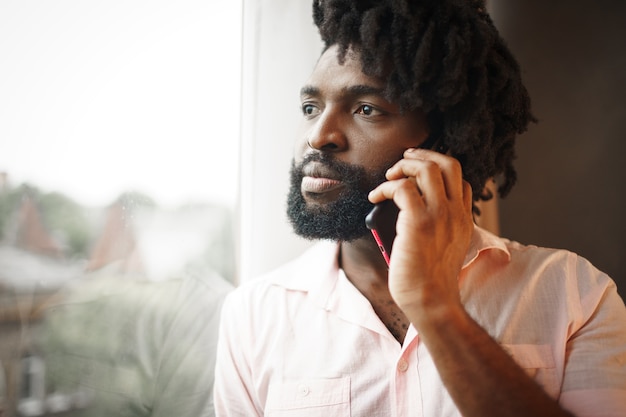 Schließen Sie herauf Foto eines schwarzen Mannes mittleren Alters im formellen Hemd, das auf seinem Telefon nahe dem Fenster spricht