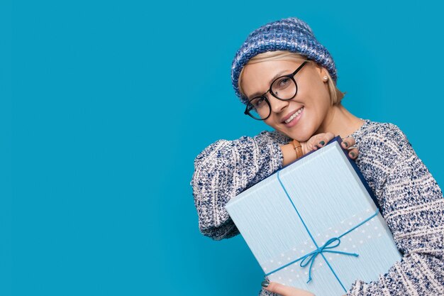 Schließen Sie herauf Foto von einer kaukasischen Frau mit Brille und Hut, die mit einem Geschenk nahe blauem Studio freiem Raum aufwirft