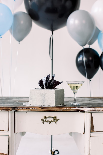 Schließen Sie herauf Foto von Geburtstagstorte und Martini-Cocktail auf dem Holztisch. Große bunte Luftballons auf dem Hintergrund. Geburtstagsfeier-Konzept