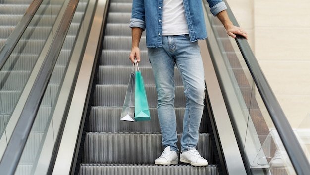 Schließen Sie herauf Foto von männlichen Beinen in Jeans mit Einkaufstüten, während er auf der Rolltreppe im Einkaufszentrum steht