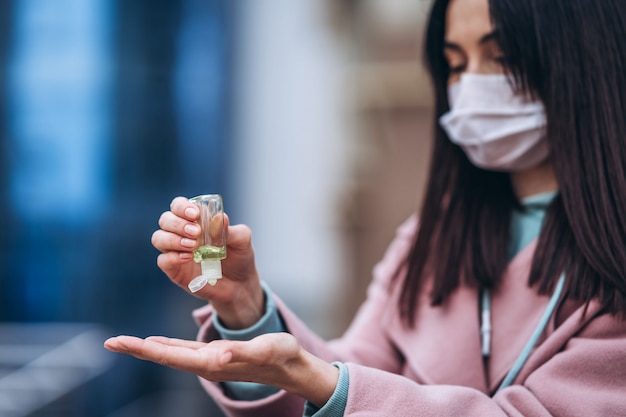 Schließen Sie herauf Hände der Frau in der medizinischen Maske, die ihre Hände mit Desinfektionsmittel im Freien in der Stadt reinigt, um Viruskrankheiten zu verhindern. Coronavirus, COVID-19, Epidemie, Pandemie, Quarantänekonzept.