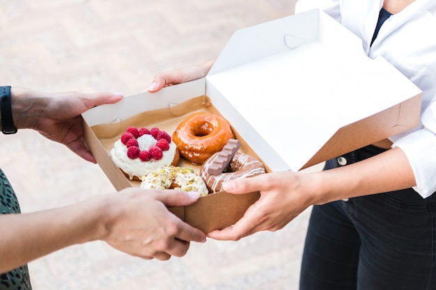 Schließen Sie herauf Hände, die vier Stücke von völlig verschiedenen bunten und köstlich aussehenden Donuts im ökologischen Karton halten