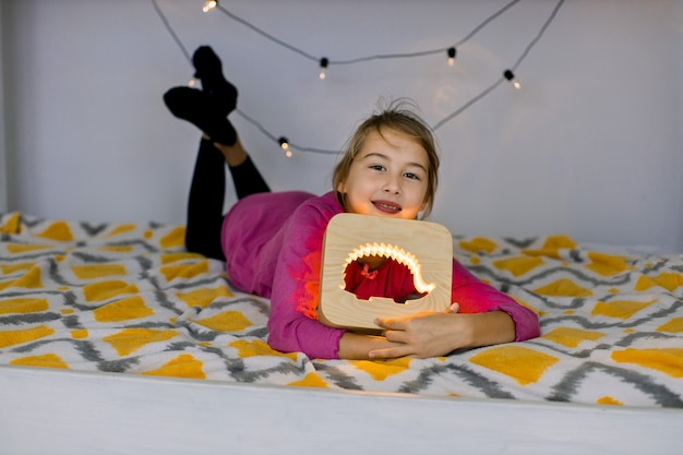 Schließen Sie herauf horizontale Aufnahme des glücklichen lächelnden 10-jährigen Mädchens, das auf gemütlichem Bett liegt und schöne hölzerne Nachtlampe mit Igelbild hält.