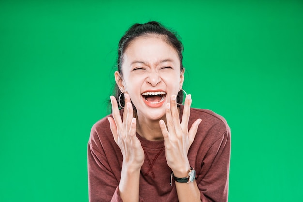 Schließen Sie herauf junge asiatische Frau, die weit geöffneten Mund ausdrückt und Hände im Staunen lokalisiert auf grünem Hintergrund anhebt