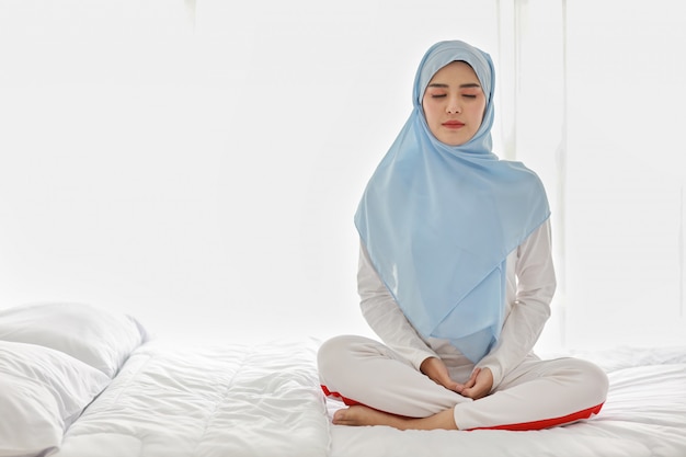 Schließen Sie herauf junge asiatische muslimische Frau, die auf Bett sitzt und Meditation genießt. Schöne Frau in Nachtwäsche mit blauem Hijab praktiziert Yoga im Schlafzimmer mit Frieden und Ruhe. Gesundes und Lifestyle-Konzept