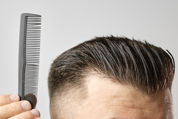 Schließen Sie herauf junger Mann, der seine Haare mit einem Plastikkamm kämmt. Haarstyling nach dem Friseursalon.