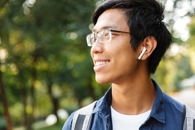 Schließen Sie herauf lächelnder asiatischer männlicher Student in Brillen und Kopfhörern, die Musik hören, während sie wegschauen