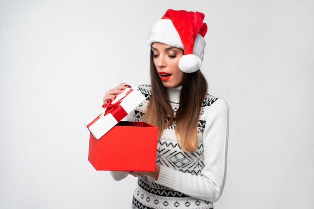 Schließen Sie herauf Porträt beautifiul kaukasische Frau in rotem Sankt-Hut auf weißer Wand. Konzept des Weihnachtsneuen Jahres überraschte nette Frauenzähne, die positive Gefühle lächeln, öffnen große rote Geschenkbox