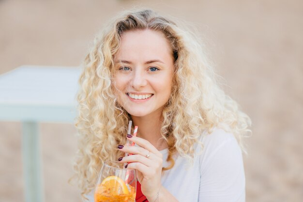 Schließen Sie herauf Porträt der attraktiven jungen Frau mit positivem Ausdruck, zeigt weiße perfekte Zähne, trinkt frisches Cocktail, wirft gegen Strand auf, hat Spaß mit Freunden, genießt Sommerzeit