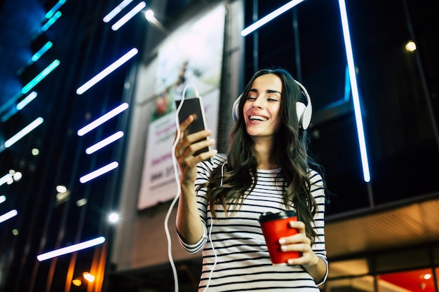 Schließen Sie herauf Porträt der attraktiven jungen glücklichen Frau in den Kopfhörern mit intelligentem Telefon in den Händen draußen