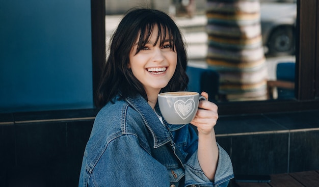 Schließen Sie herauf Porträt der Dame mit den schwarzen Haaren, die in die Kamera lächeln und eine Tasse Kaffee trinken