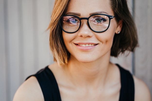 Schließen Sie herauf Porträt der jungen glücklichen Frau in den Gläsern mit hübschem Lächeln und blauen Augen, kurzen Haaren, lässig gekleidet. Konzept der glücklichen Leute.