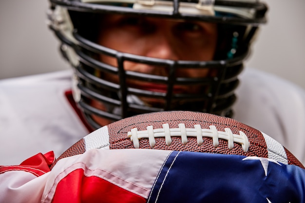 Foto schließen sie herauf porträt des amerikanischen fußballspielers im helm mit ball und amerikanischer flagge stolz auf sein land, auf einem weißen raum.