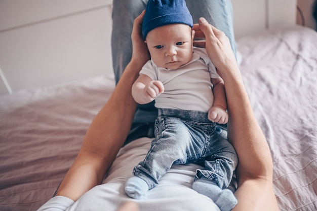 Schließen Sie herauf Porträt des glücklichen jungen Vatervaters, der sein Baby in den blauen Jeans und im weißen T-Shirt und in der Kappe hält. Junge glückliche Familie, Vater spielt mit niedlichen emotionalen kleinen neugeborenen Kind Sohn im Schlafzimmer.