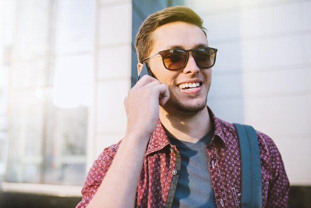 Schließen Sie herauf Porträt des netten attraktiven Lächelns kaukasischer Mann in der stilvollen Sonnenbrille in der Stadt Junger Geschäftsmann, der auf intelligentem Telefon auf dem Gebäude im Freien spricht Mode-urbaner Lifestyle-Konzept Reisender