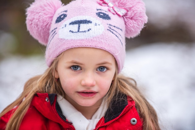 Schließen Sie herauf Porträt des schönen blonden Mädchens, das in der warmen Wintermütze auf einem Schneehintergrund spielt. Nettes Kindergesicht.