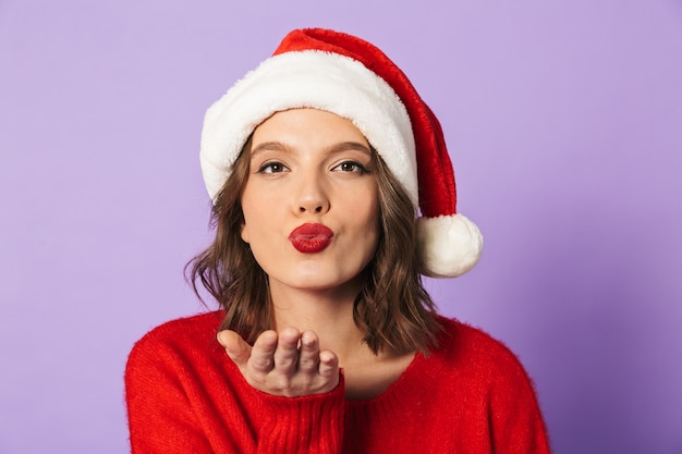 Schließen Sie herauf Porträt einer hübschen jungen Frau, die roten Weihnachtshut trägt, der über violetter Wand lokalisiert wird