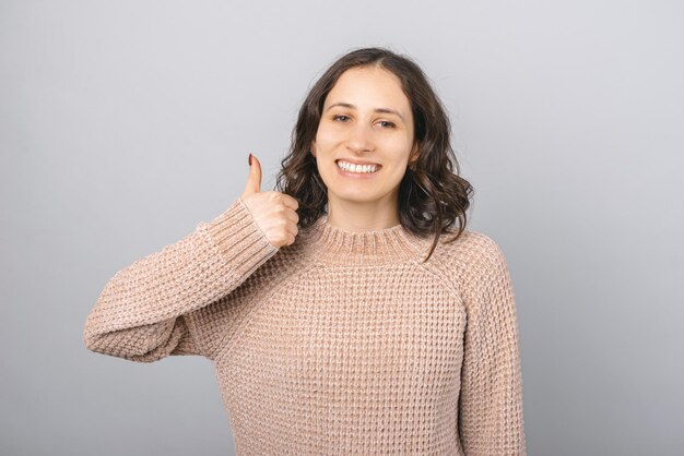 Schließen Sie herauf Porträt einer jungen glücklichen Frau mit den kurzen Haaren, die wie Geste Studioaufnahme über grauem Hintergrund zeigen