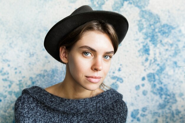 Schließen Sie herauf Porträt einer schönen jungen Frau, gekleidet in Pullover und Hut über blauer Wand