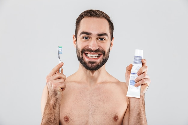 Schließen Sie herauf Porträt eines hübschen bärtigen Mannes, der isoliert über Weiß steht und Zahnpasta und Zahnbürste zeigt