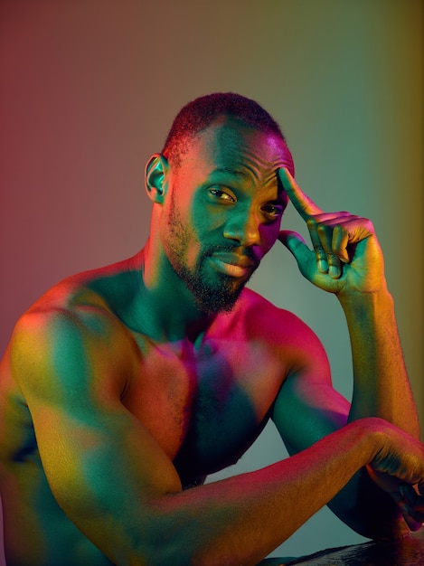 Schließen Sie herauf Porträt eines jungen nackten traurigen afrikanischen Mannes, der Kamera betrachtet. High Fashion männliches Model in bunten hellen Lichtern posiert im Studio. Kunstdesign über lebendigem Hintergrund.