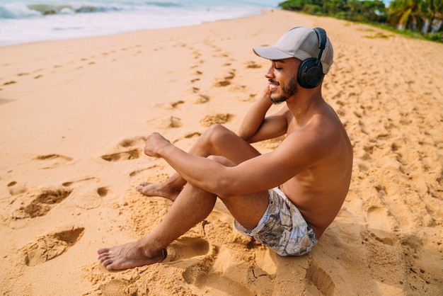 Schließen Sie herauf Porträt eines lateinamerikanischen Mannes, der auf dem Sand sitzt, der Musik hört