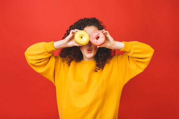 Schließen Sie herauf Porträt eines zufriedenen hübschen jungen Mädchens, das Donuts lokalisiert über rotem Hintergrund isst.