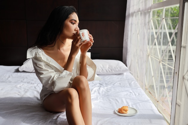 Schließen Sie herauf schöne asiatische Frau im sexy Kleid, das Kaffee und Hörnchen beim Sitzen im Bett frühstückt