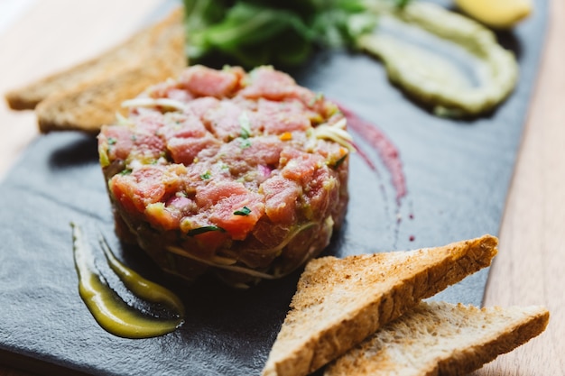 Schließen Sie herauf würzigen Bluefin-Thunfisch Tartare mit saurer und würziger Soße. Serviert mit Toast und Salat auf schwarzer Steinplatte.