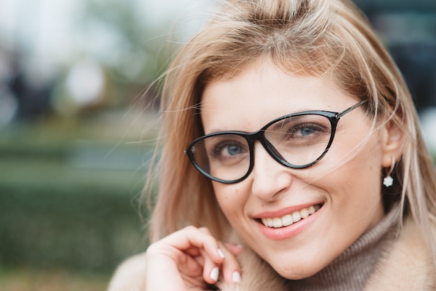 Schließen Sie oben eine hübsche junge blonde Frau in Brille und Pelzmantel im Freien lächelt und der Wind weht und spielt mit ihren Haaren im Winter