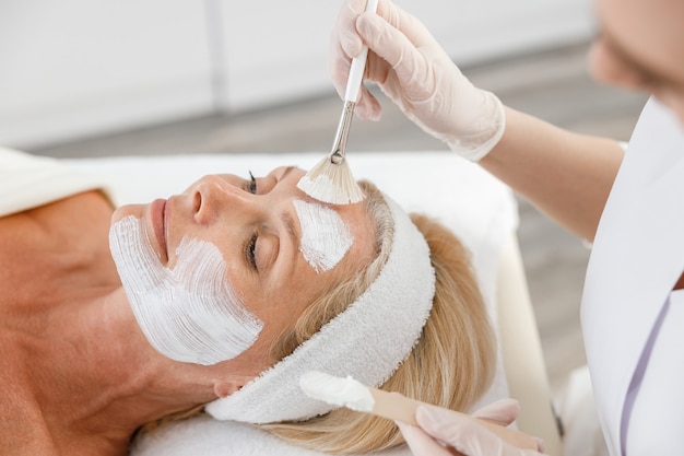 Schließen Sie oben Gesicht Peeling-Maske, Spa-Schönheitsbehandlung, Hautpflege. Ältere Frau, die Gesichtspflege durch Kosmetikerin am Spa-Salon erhält.