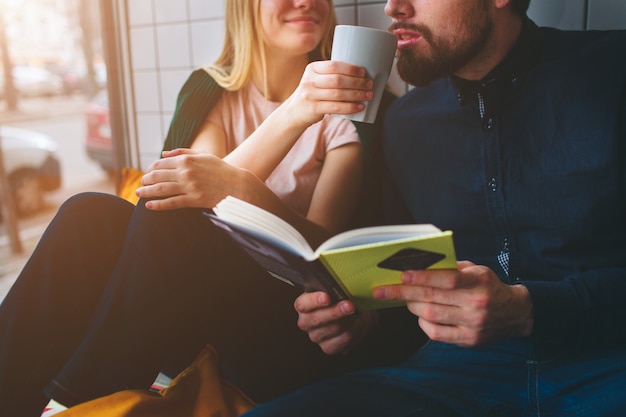 Foto schließen sie oben und schneiden sie ansicht eines jungen und des mädchens, die im café nahe großem fenster sitzen. sie trinkt kaffee und gibt ihre tasse, damit er dieses getränk probieren kann. ihr mann hält ein buch in der hand.