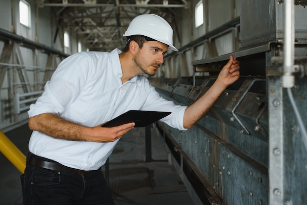 Schließen Sie oben vom jungen kaukasischen lächelnden Arbeiter mit Helm auf Kopf, der Tablette für Arbeit beim Stehen im Lager verwendet