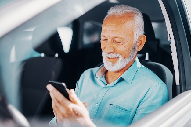 Schließen Sie oben vom lächelnden bärtigen Senior unter Verwendung des Smartphones zum Schreiben oder Lesen der Nachricht mit Arm auf dem geöffneten Fenster, während Sie im Auto sitzen.