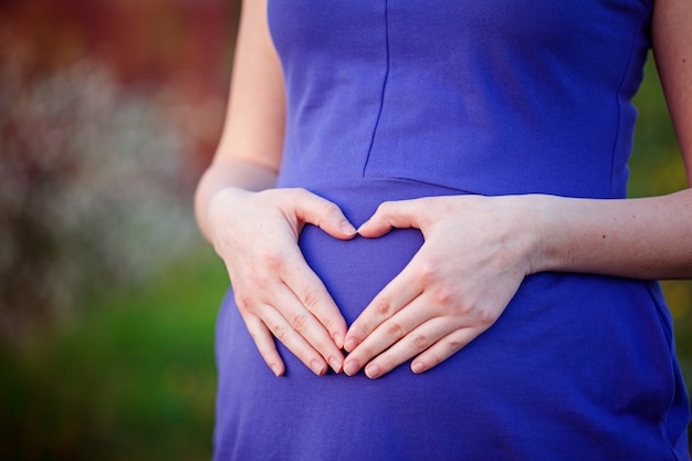 Schließen Sie oben von den Händen der jungen Frau auf dem Bauch, der die Herzform schafft, die blaues Kleid trägt.