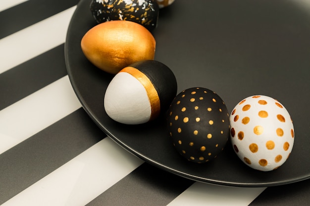 Schließen Sie oben von den schwarzen, weißen und goldenen verzierten Ostereiern auf schwarzem Teller auf gestreiftem Hintergrund. Trendy Urlaubshintergrund.